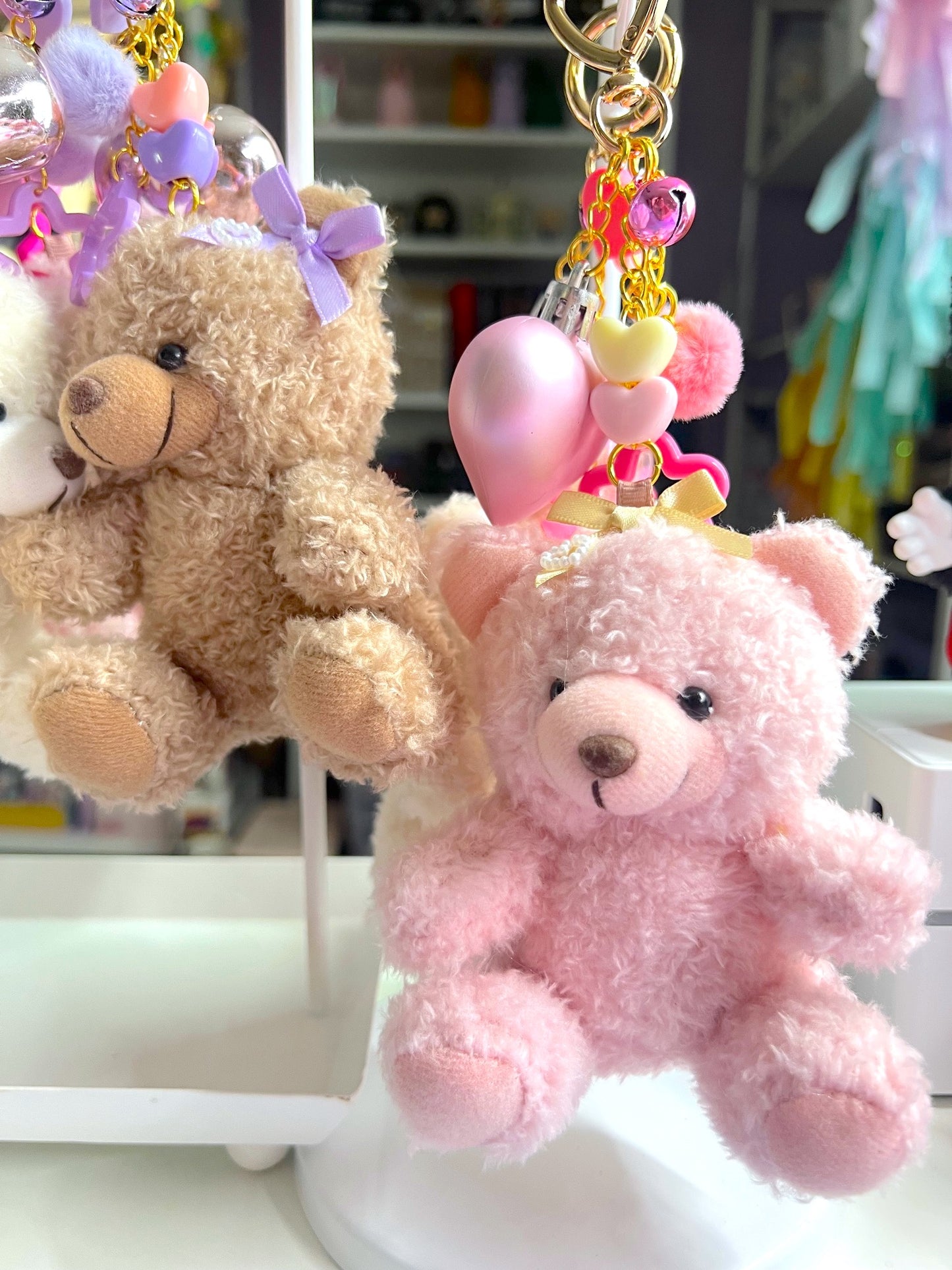 Two teddy bear keychains, one pink and one brown, with decorative elements on a blurred background.