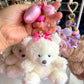 White teddy bear keychain with pink bow held by a hand against a display of other keychains.