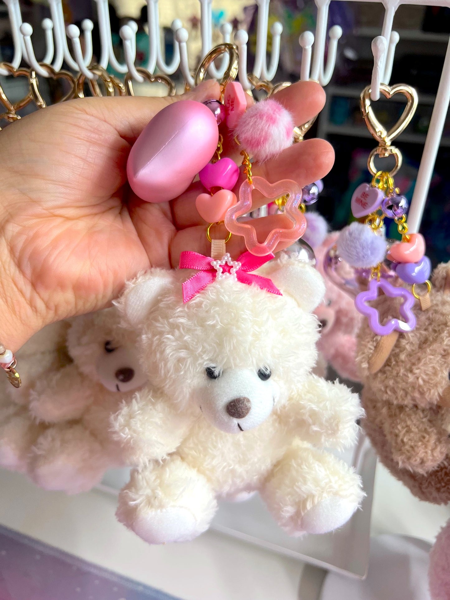 White teddy bear keychain with pink bow held by a hand against a display of other keychains.