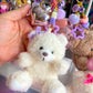 Plush teddy bear keychain with decorative elements held by a hand against a colorful background.