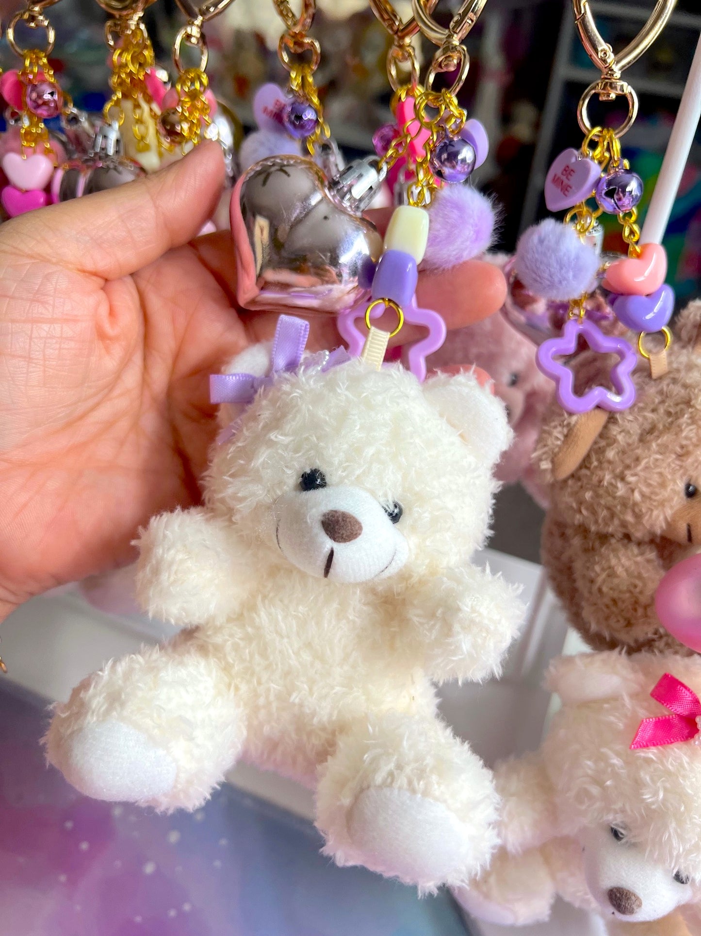 Plush teddy bear keychain with decorative elements held by a hand against a colorful background.