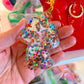 Hand holding a colorful glittery keychain with a blurred background of a red handbag and colorful objects.