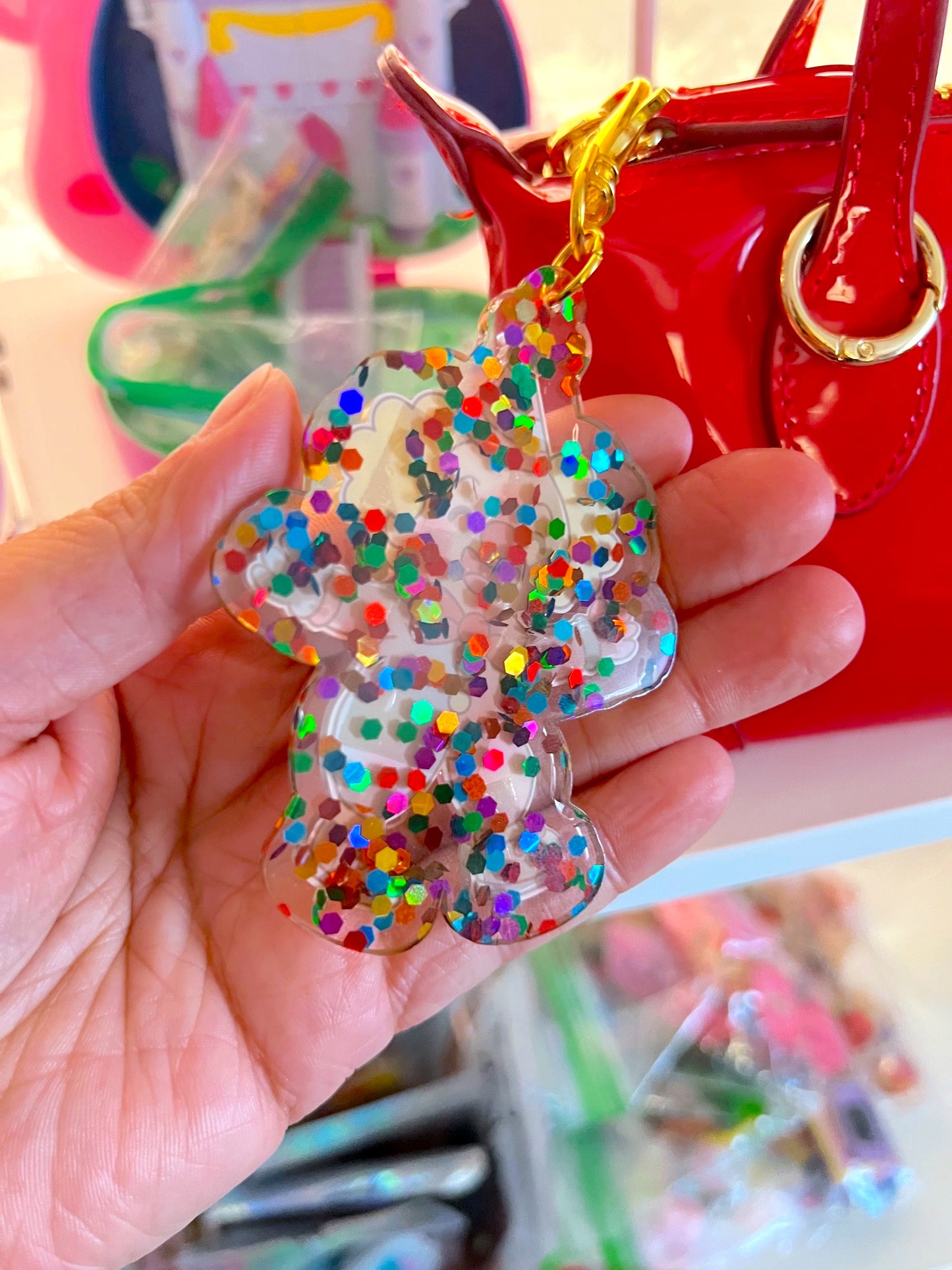 Hand holding a colorful glittery keychain with a blurred background of a red handbag and colorful objects.