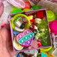 Hand holding a small box filled with colorful miniature toys and items, with a pink basket in the background.