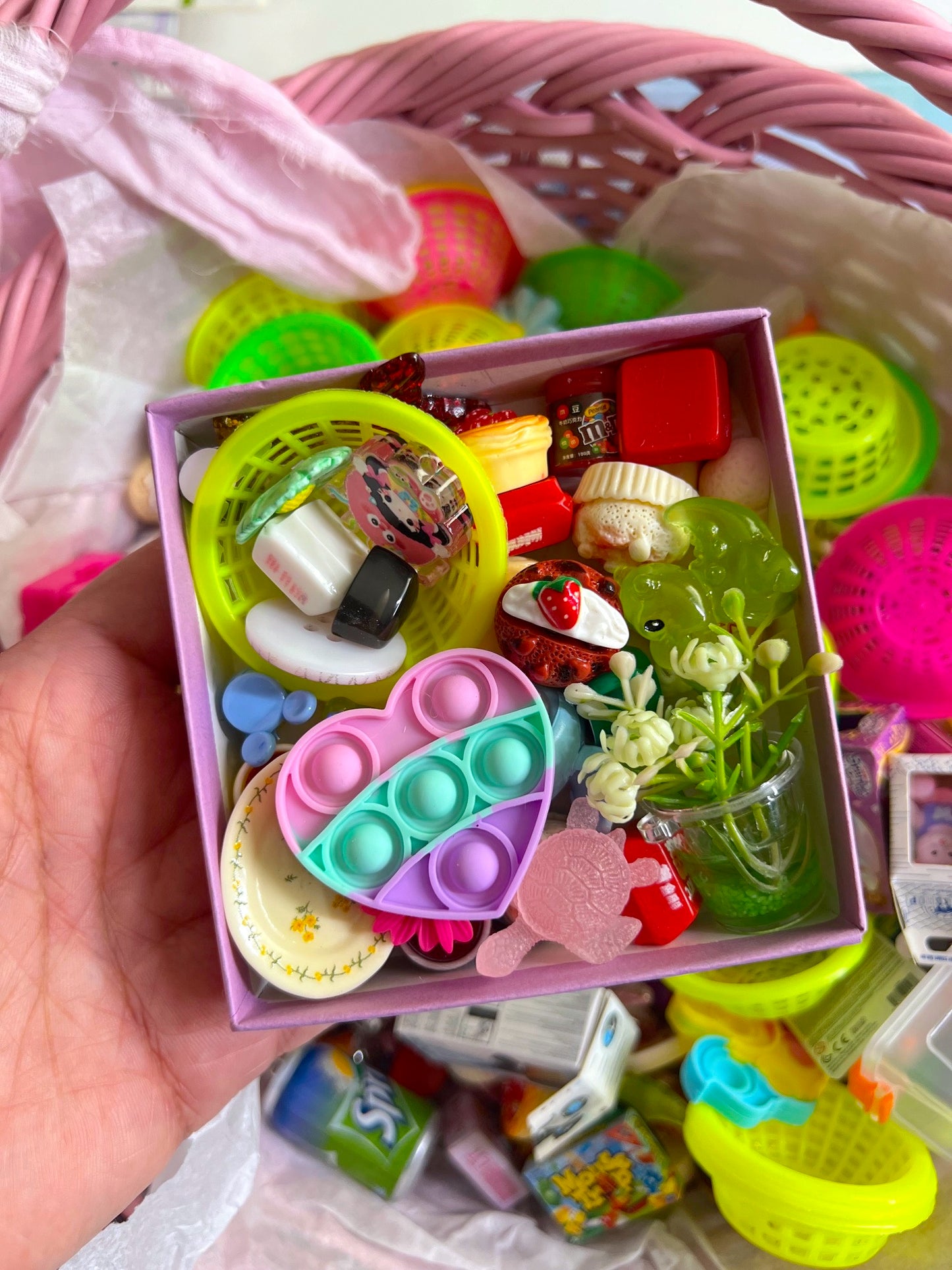Hand holding a small box filled with colorful miniature toys and items, with a pink basket in the background.