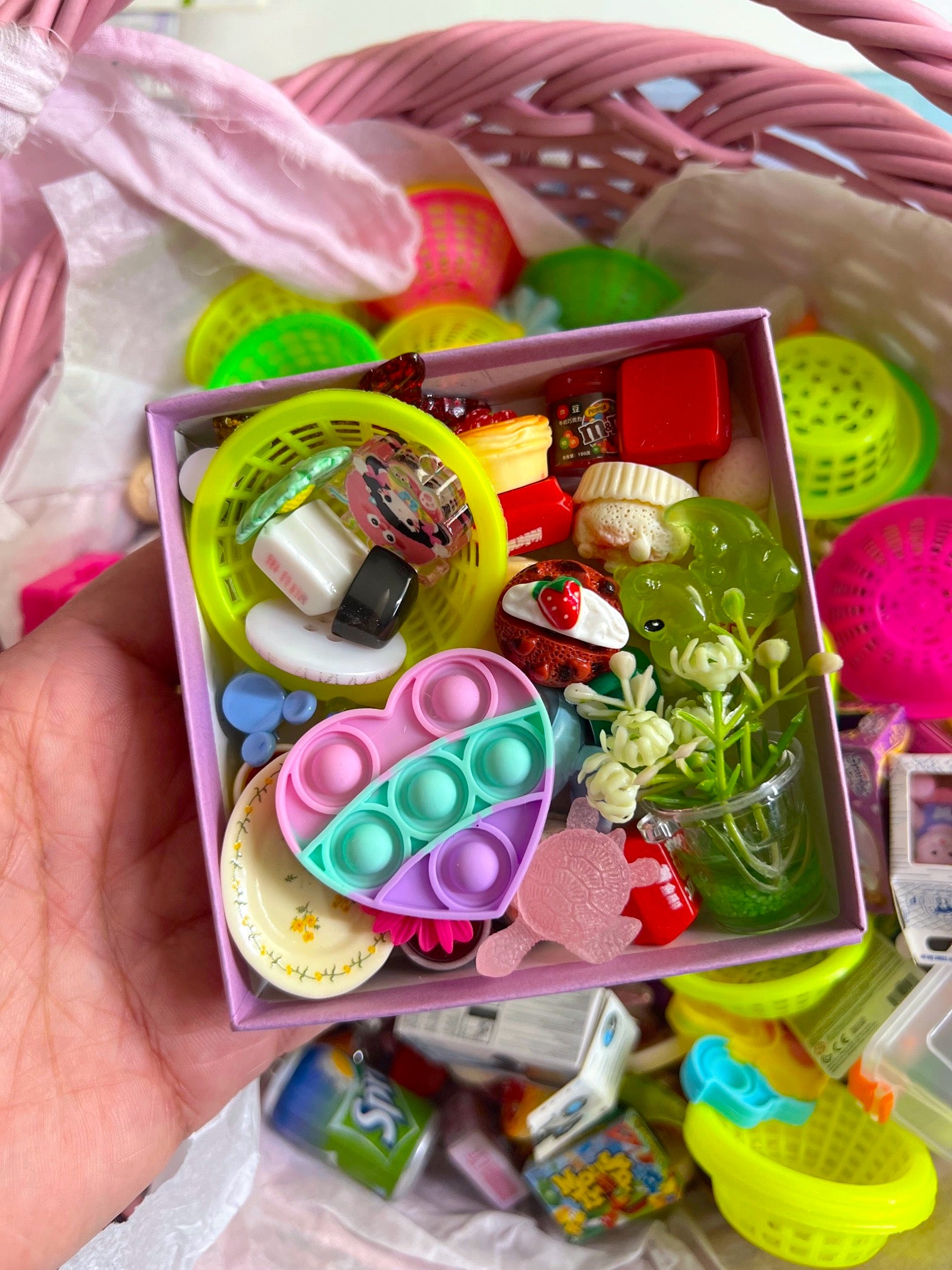 Hand holding a small box filled with colorful miniature toys and items, with a pink basket in the background.