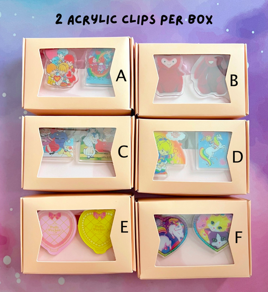 Six boxes containing acrylic clips with colorful designs on a purple background.